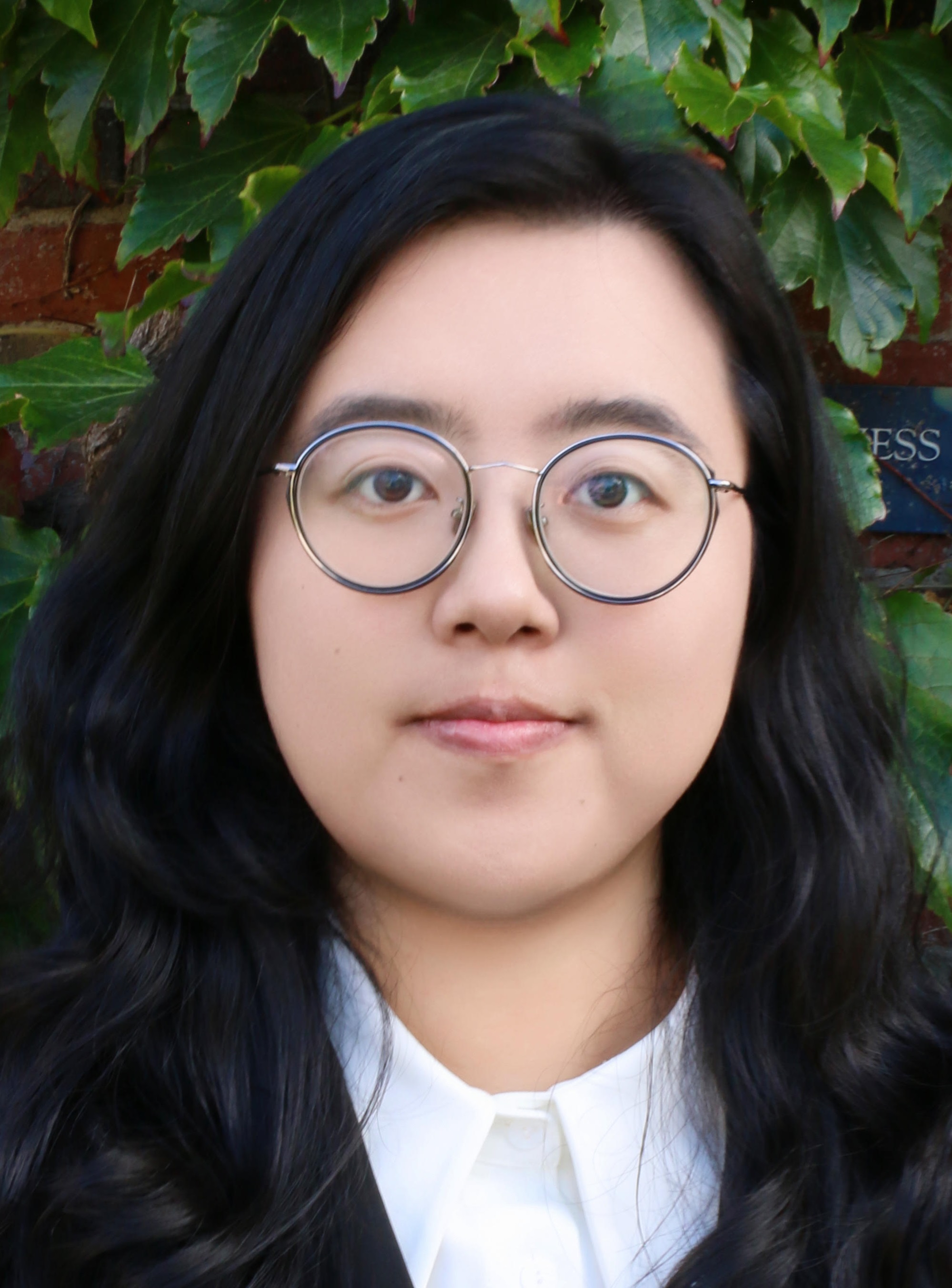 Close-up portrait of a person with shoulder-length black hair and round glasses, standing in front of a vine-covered brick wall.