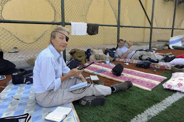 Marie Colvin sits on the ground in a fenced-in refugee camp surrounded by refugees. She is holding a tape recorder and has a notebook on her lap.