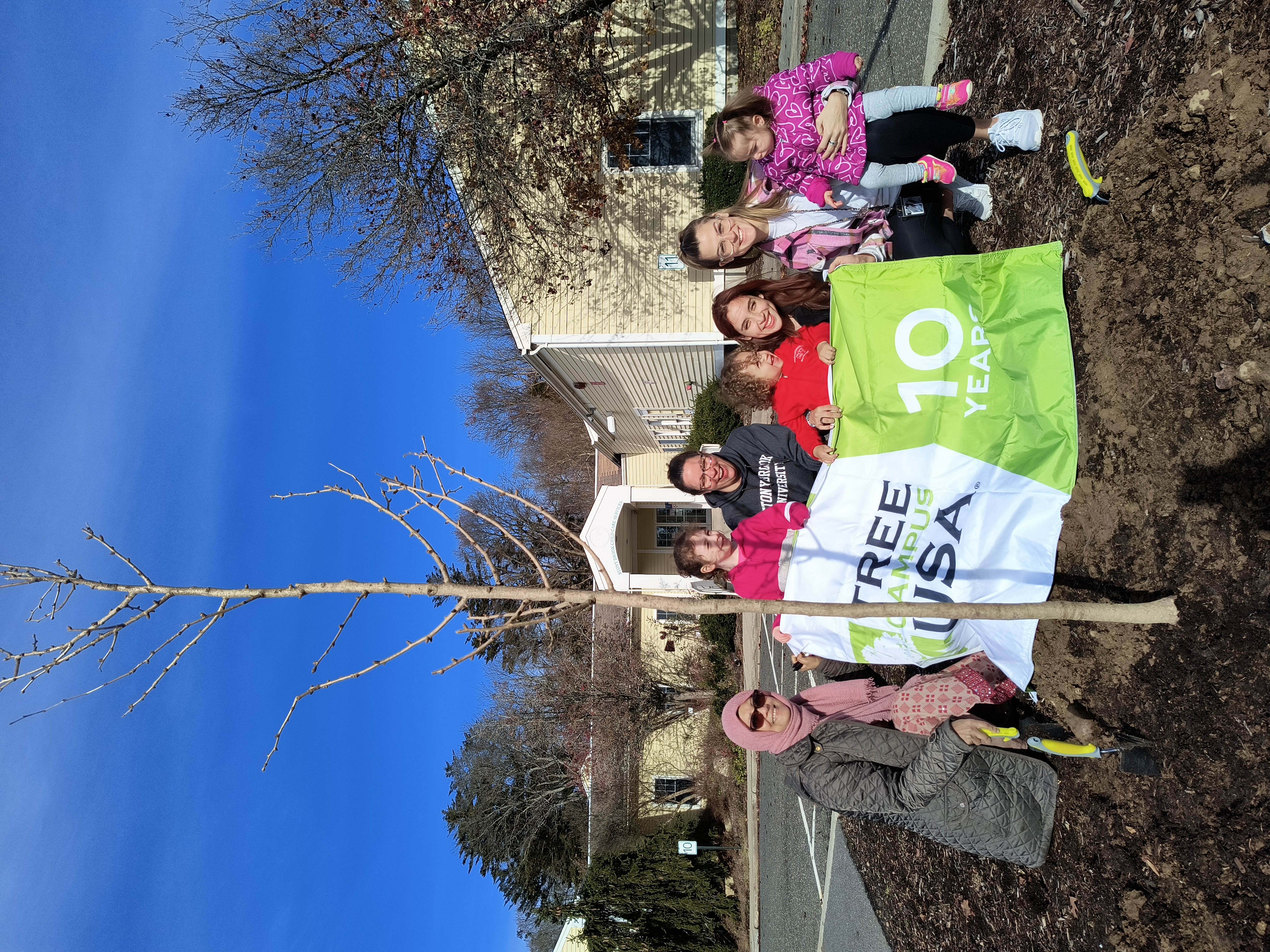 image of adults and children standing behind a newly planted tree in front of the stony brook child care services center