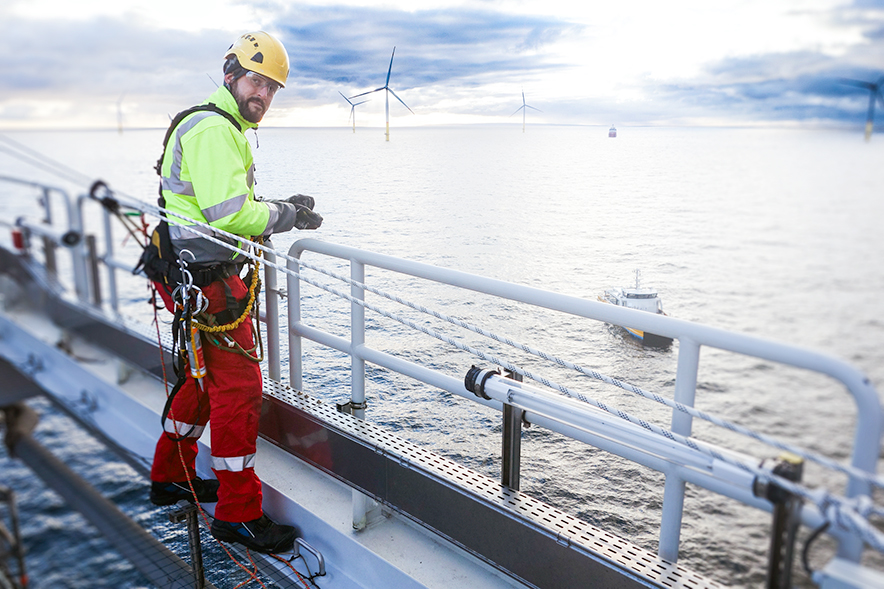 Man with a hardhat and harness is standing on a platform over the sea, wind turbines in the distance